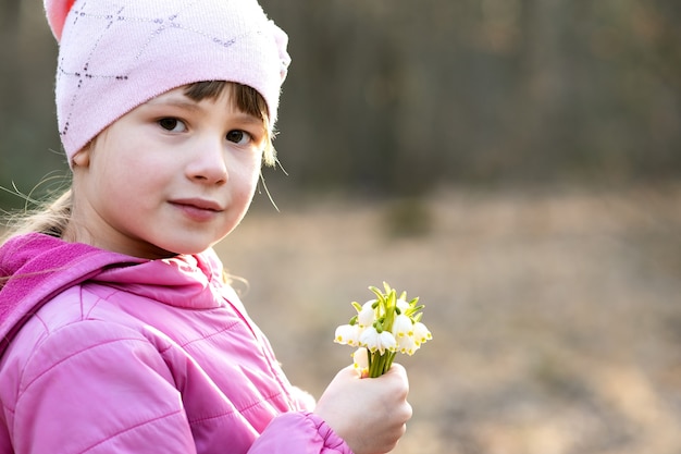 Porträt des glücklichen Kindermädchens, das Bündel der frühen Frühlingsschneeglöckchenblumen draußen hält.