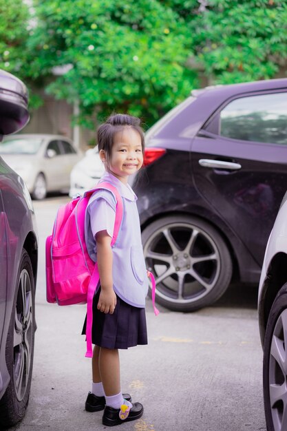Porträt des glücklichen kleinen Mädchens in der thailändischen Schuluniform mit dem Rucksack, der im Parkplatz steht