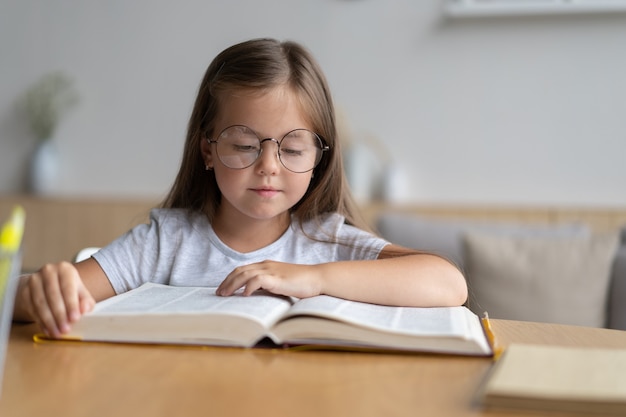 Porträt des glücklichen kleinen Schülers, der zu Hause lernt. Lächelndes kleines Kindermädchen, das den Unterricht im Wohnzimmer genießt. Intelligentes Kinderschulmädchen, das in die Kamera schaut