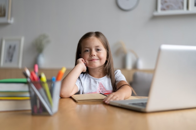 Porträt des glücklichen kleinen Schülers, der zu Hause lernt. Lächelndes kleines Kindermädchen, das den Unterricht im Wohnzimmer genießt. Intelligentes Schulmädchen, das in die Kamera schaut und aus der Ferne online lernt.