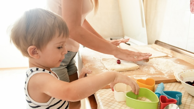 Porträt des glücklichen lächelnden Kleinkindjungen mit der jungen Mutter, die auf Küche backt und kocht. Eltern unterrichten und erziehen das Kind zu Hause