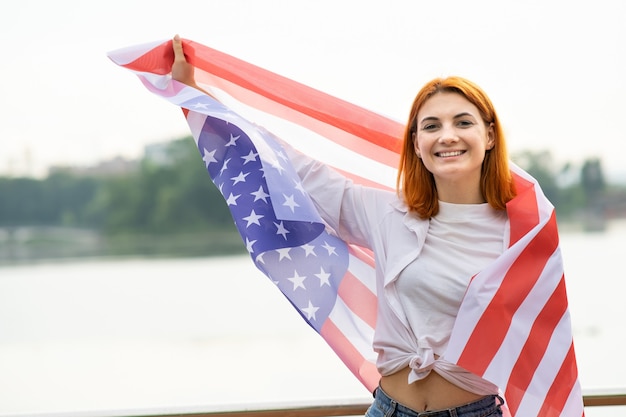 Porträt des glücklichen lächelnden rothaarigen Mädchens mit USA-Nationalflagge in ihren Händen