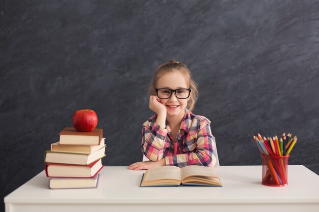 Porträt des glücklichen netten intelligenten lächelnden Mädchens in den Gläsern, die mit Stapel Büchern am Tisch sitzen, Kopienraum. Bildungs- und Entwicklungskonzept