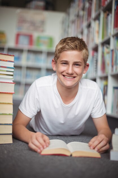 Foto porträt des glücklichen schuljungen, der auf boden mit büchern in der bibliothek liegt