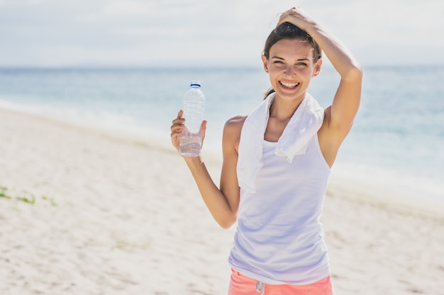 Porträt des glücklichen sportlichen Mädchens, das eine Flasche Mineralwasser zur Erfrischung am Strand hält