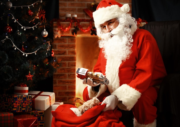 Porträt des glücklichen Weihnachtsmanns, der in seinem Zimmer zu Hause in der Nähe von Weihnachtsbaum und großem Sack sitzt und Weihnachtsbrief oder Wunschliste liest