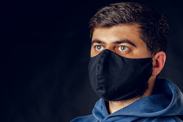 Porträt des gutaussehenden Mannes, der medizinische schwarze Maske auf dem Gesicht während der Virusepidemiesperre trägt, die auf dunkler Wand aufwirft. Nahaufnahme.