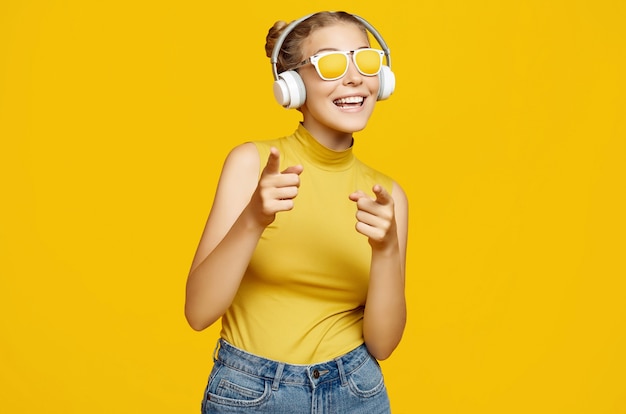 Porträt des herrlichen blonden Hipster-Mädchens mit dem lockigen Haar, das Sonnenbrille trägt, genießt die Musik in den Kopfhörern auf gelbem Hintergrund