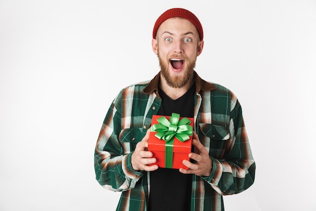 Porträt des hübschen bärtigen Kerls, der Hut und kariertes Hemd hält Geschenkgeschenk hält, während lokalisiert über weißem Hintergrund steht