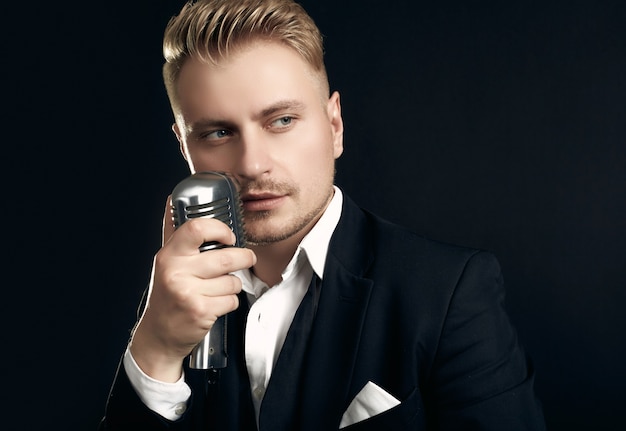 Porträt des hübschen blonden Mannesängers im eleganten Smoking und in der Fliege, die mit Weinlesemikrofon auf schwarzer Wand aufwirft
