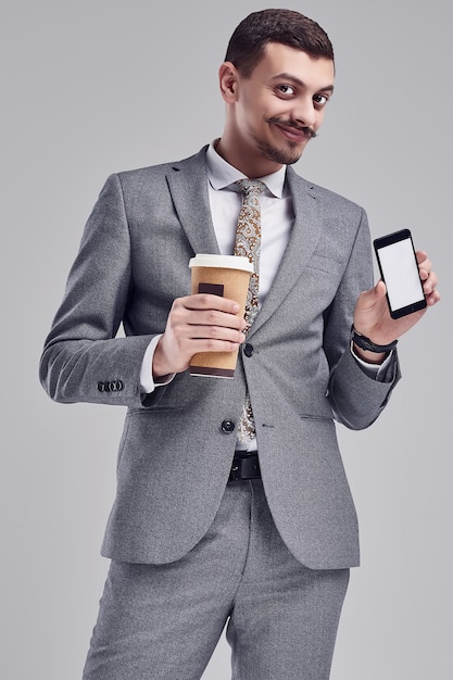 Porträt des hübschen jungen überzeugten arabischen Geschäftsmannes mit grauem vollem Anzug des fantastischen Schnurrbartes in Mode hält einen Tasse Kaffee und ein Telefon auf Studio