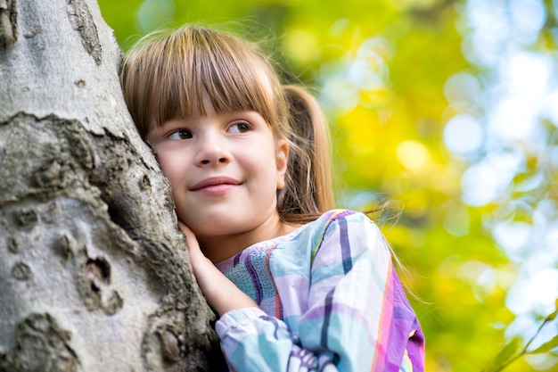 Porträt des hübschen Kindermädchens, das sich an einen Baumstamm im Herbstpark lehnt, der sich entspannt. Nettes weibliches Kind, das warmes Herbstwetter im Wald genießt.