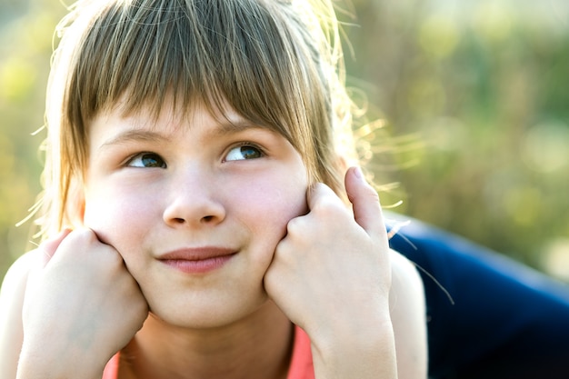 Porträt des hübschen Kindermädchens mit grauen Augen und langen blonden Haaren, die sich auf ihre Hände stützen und glücklich draußen auf verschwommener heller Oberfläche lächeln