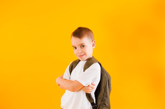 Porträt des hübschen kleinen Jungen steht mit einem Rucksack auf gelbem Hintergrund Der Typ in einem weißen T-Shirt steht mit verschränkten Armen