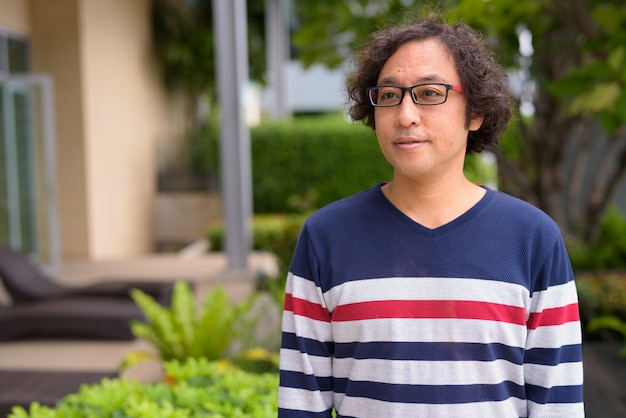 Porträt des japanischen Mannes mit dem lockigen Haar, das Brillen mit Natur im Dachgarten draußen trägt