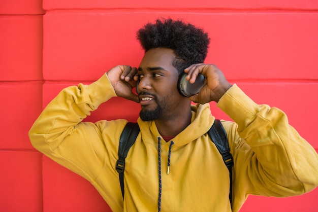 Porträt des jungen Afro-Mannes, der Musik mit Kopfhörern gegen roten Raum hört. Technologiekonzept.