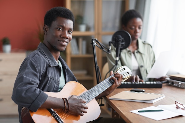 Porträt des jungen afroamerikanischen Mannes, der akustische Gitarre spielt und Kamera betrachtet, während Musik im Hauptaufnahmestudio komponiert, Raum kopiert