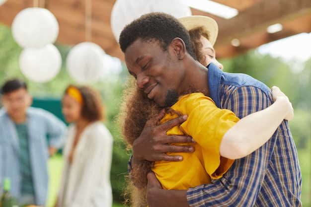 Porträt des jungen afroamerikanischen Mannes, der Freund liebevoll umarmt, während sie sich auf der Party im Freien im Sommer begrüßen