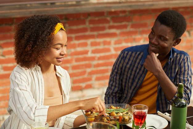 Porträt des jungen afroamerikanischen Mannes, der schöne Frau beim Genießen des Abendessens mit Freunden im Freien am Tisch während der Sommerparty betrachtet