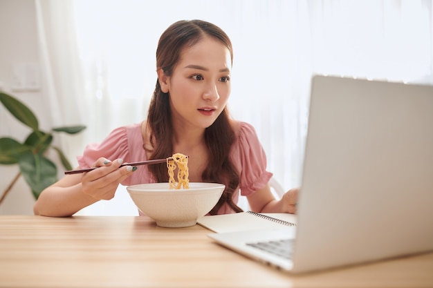 Porträt des jungen asiatischen Mädchens, das Laptop beim Essen von Nudeln zu Hause verwendet.