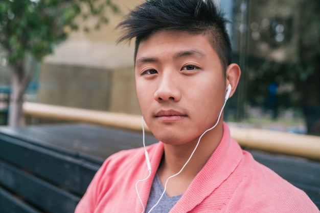 Porträt des jungen asiatischen Mannes, der Musik mit Kopfhörern beim Sitzen auf einer Bank in einem Park hört.