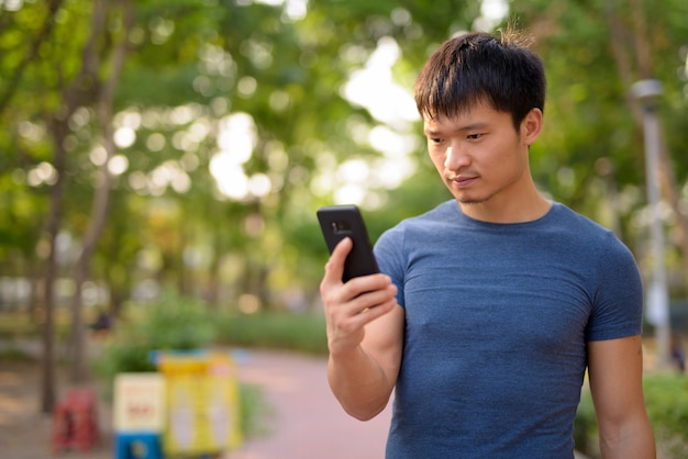 Porträt des jungen asiatischen Mannes, der Telefon am Park im Freien verwendet