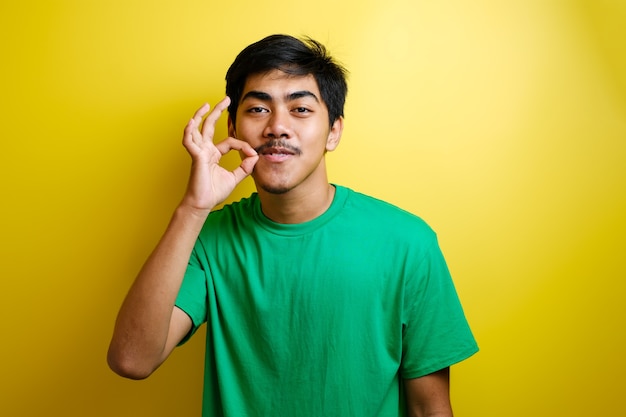 Porträt des jungen asiatischen Mannes im grünen T-Shirt, das köstliche Handgeste vor gelbem Hintergrund macht