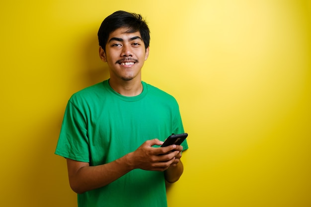 Porträt des jungen asiatischen Mannes Lächeln Blick in die Kamera beim Surfen im Internet auf dem Smartphone, vor gelbem Hintergrund Studioaufnahme