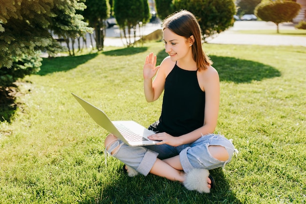 Porträt des jungen attraktiven Mädchens, das online per Videoanruf kommuniziert, Skype-Gespräch mit Freunden führt, einen Laptop benutzt, während auf Gras im Park sitzt.