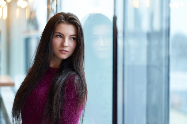 Porträt des jungen Brunettemädchens, das nahes Fenster steht