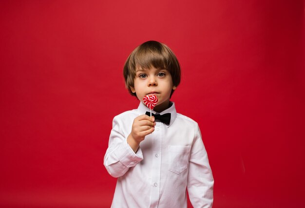 Porträt des Jungen, der einen Lutscher in der Form eines Herzens auf einem roten Hintergrund mit einer Kopie des Raumes hält