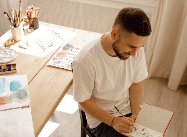 Porträt des jungen dunkelhaarigen konzentrierten lächelnden kaukasischen weißen männlichen Künstlers mit einem Bart in einem weißen T-Shirt im Studio, das durch Sonnenlicht beleuchtet wird. in einem Studio in Holztisch sitzen.