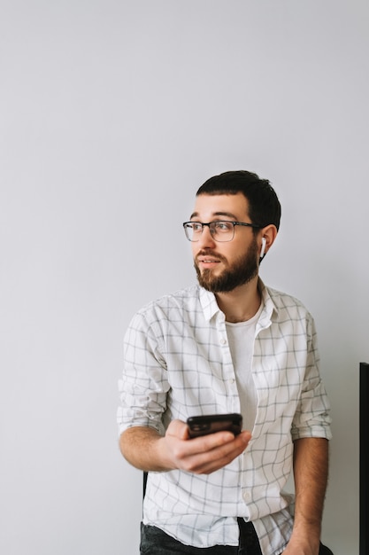 Porträt des jungen fröhlichen kaukasischen Mannes in den Gläsern und in den Kopfhörern unter Verwendung des Mobiltelefons.