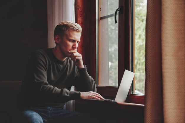 Porträt des jungen Geschäftsmannes mit Laptop am Fenster des Wohnzimmers im Landhaus. Workaholic-Mann in Freizeitkleidung zu Hause, die im Urlaub arbeitet. Kreative Inspiration und Start-up-Business. Platz kopieren