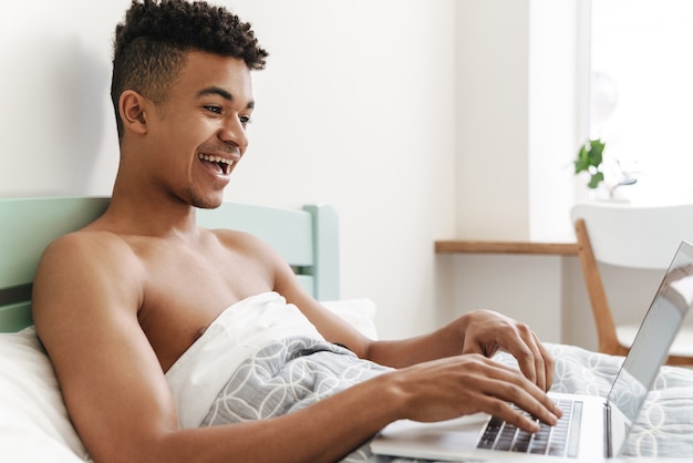 Porträt des jungen glücklichen afrikanischen Kerls morgens im Schlafzimmer mit Laptop-Computer im Bett.