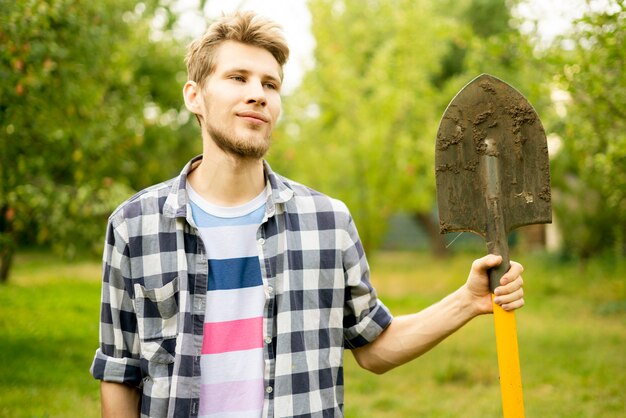 Porträt des jungen glücklichen Bauern, der Schaufel auf einer Sommerlandwirtschaftsökofarm a trägt