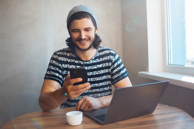 Porträt des jungen glücklichen Mannes, der Smartphone verwendet und an seinem Laptop mit Kaffeetasse auf Schreibtisch arbeitet.