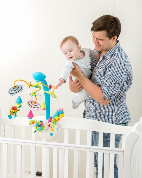 Porträt des jungen glücklichen Vaters, der seinem kleinen Sohn ein neues Spielzeugkarussell im Kinderbett zeigt