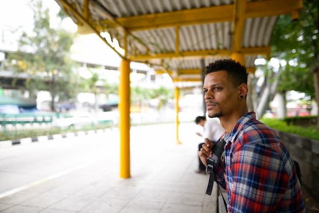 Porträt des jungen gutaussehenden bärtigen afrikanischen Hipster-Mannes als Tourist mit Rucksack, der an der Bushaltestelle in der Stadt draußen wartet