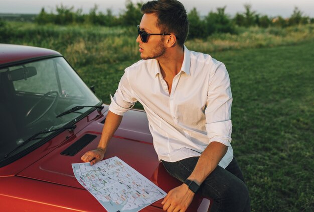 Porträt des jungen gutaussehenden Mannes, der eine Karte auf einer Autoreise für Reiseführerziel im Urlaub verwendet Kaukasischer Geschäftsmann, der auf einer Karte schaut, während er auf der Motorhaube des Autos auf Naturhintergrund sitzt Reisen