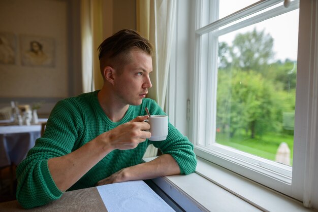 Porträt des jungen gutaussehenden Mannes, der Kaffee trinkt und zu Hause aus dem Fenster schaut