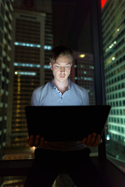 Porträt des jungen gutaussehenden Mannes, der Laptop gegen Glasfenster mit Blick auf die Stadt bei Nacht verwendet