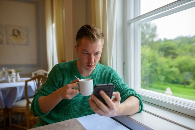 Porträt des jungen gutaussehenden Mannes, der Telefon benutzt und Kaffee am Fenster zu Hause trinkt