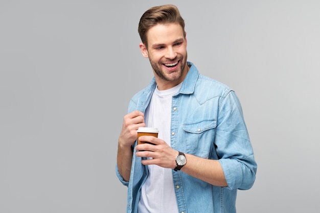 Porträt des jungen gutaussehenden Mannes im Jeanshemd über der hellen Wand, die Tasse Kaffee hält, um zu gehen