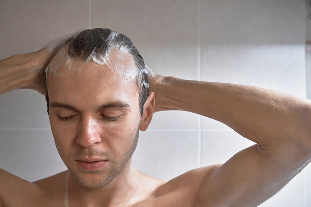 Porträt des jungen gutaussehenden Mannes wäscht sich mit Duschgel, schäumt Kopf mit Shampoo im Badezimmer zu Hause Nahaufnahme