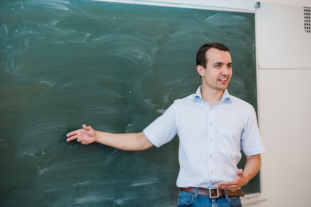 Porträt des jungen gutaussehenden Schülers oder Lehrers in einer Klasse, die auf leere Tafel zeigt, spricht und lächelt