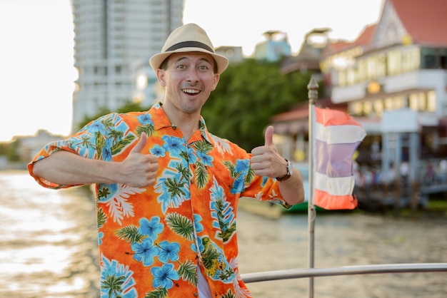 Porträt des jungen gutaussehenden Touristenmannes am Pier auf der Ansicht des Flusses in der Stadt Bangkok