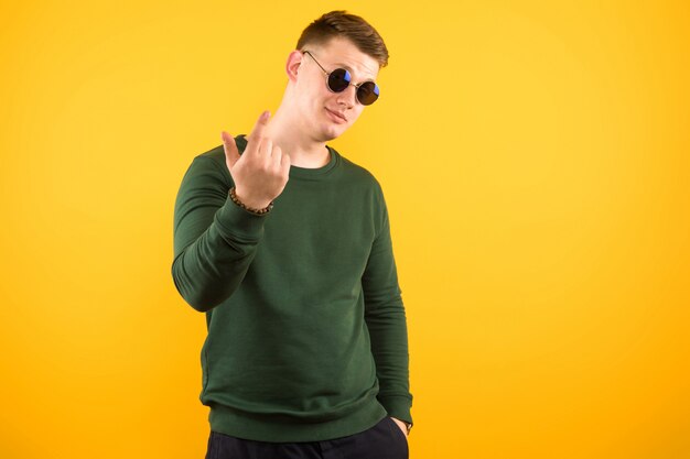 Porträt des jungen hübschen lächelnden Kerls mit der Sonnenbrille, die Geste auf gelbem Hintergrund zeigt