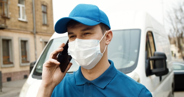 Porträt des jungen Kuriers in der Maske, die nahe Auto hält, das Kartonbox unter Verwendung des Smartphones hält