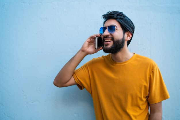 Porträt des jungen lateinischen Mannes, der am Telefon gegen blauen Hintergrund spricht. Kommunikationskonzept.